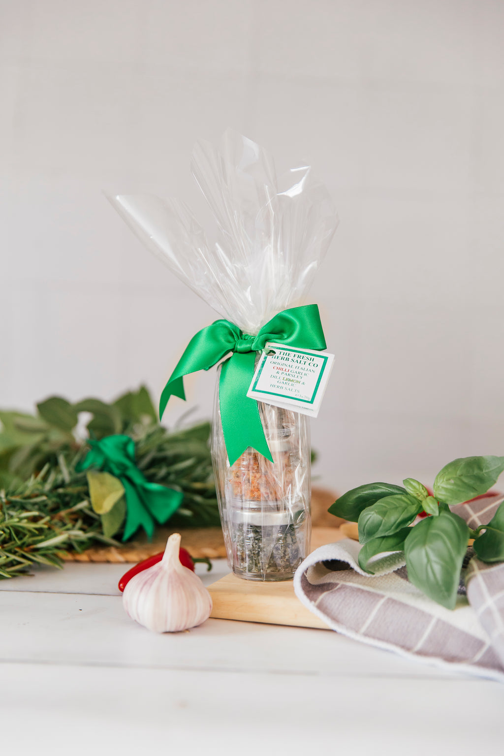 A visually appealing Gift Pack Trio of Fresh Herb Sea Salt Seasonings, elegantly presented in cellophane wrap and adorned with a vibrant green bow. The pack includes three 20g pots of delectable flavours: Original Italian with rosemary, basil, sage, garlic, and bay; Chilli Garlic & Parsley; and Herbes de Provence with marjoram, sage, parsley, and thyme. A perfect culinary gift ensemble with a touch of sophistication.