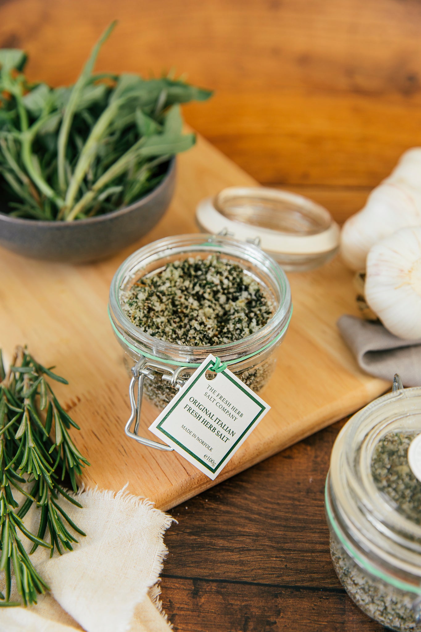 Aromatic and visually stunning, this image depicts a glass Kilner jar containing Original Italian Fresh Herb Sea Salt Seasoning. The jar showcases a flavourful mix of rosemary, garlic, basil, sage and bay, mixed into pristine blend of white coarse and fine sea salt in a carefully arranged kitchen scene.
